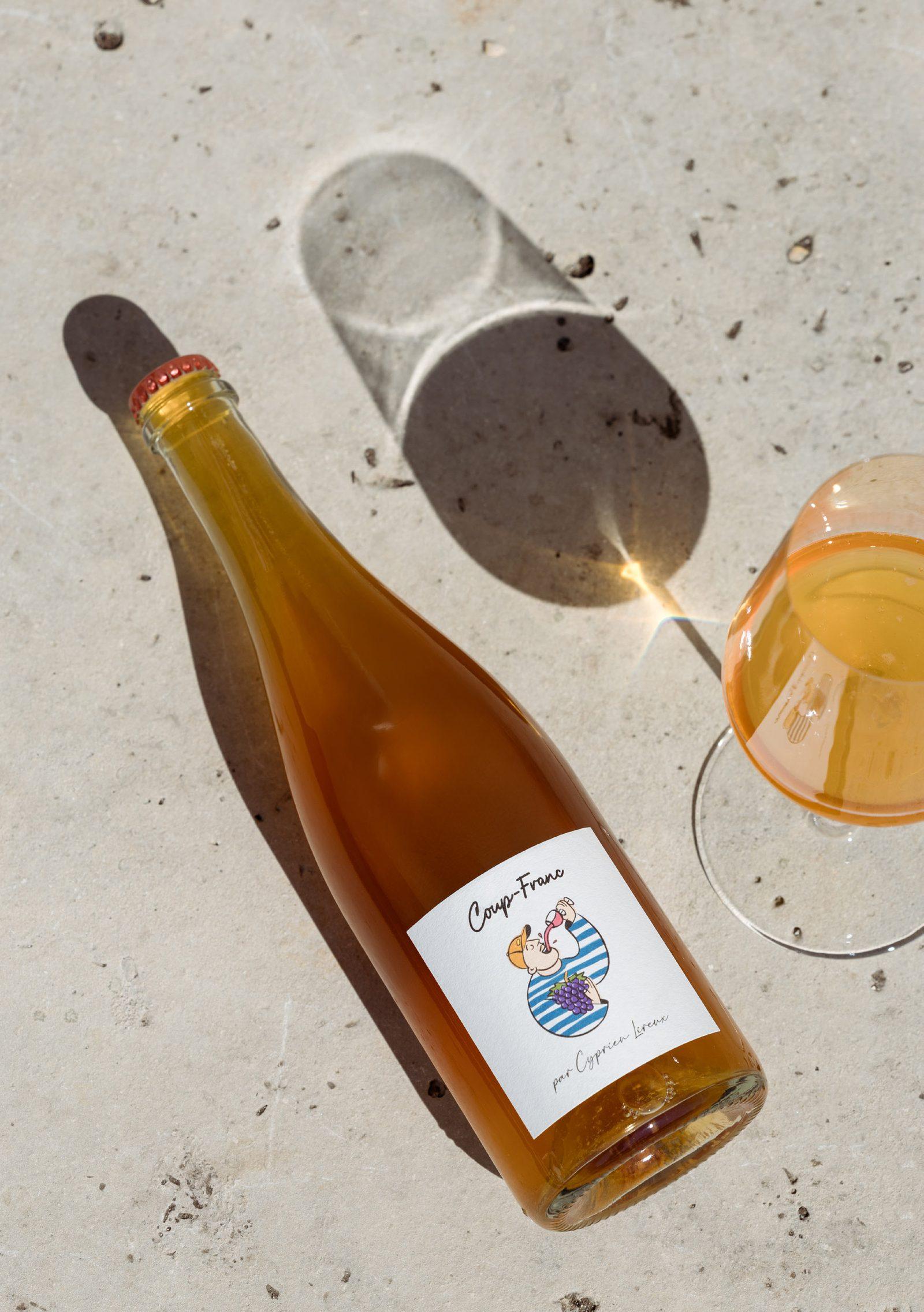 A bottle of Coup-Franc lying on sunlit pale stone beside a glass of the amber co-ferment, shadows falling long