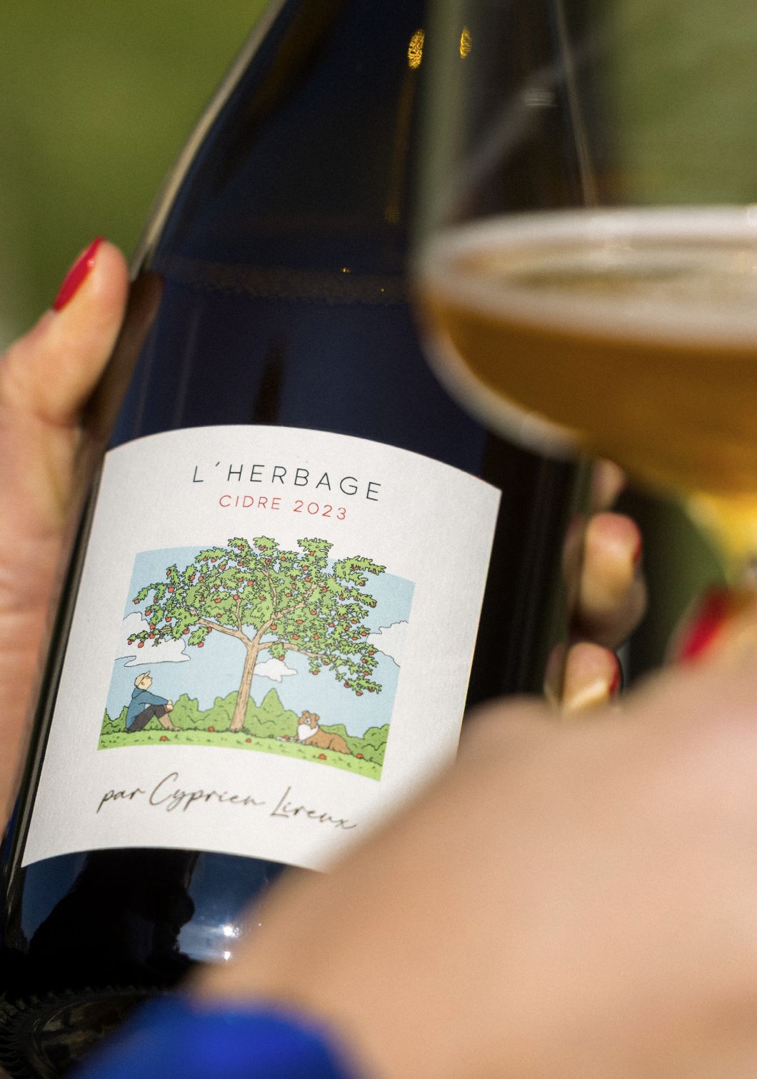 A bottle of L'Herbage cidre held up close, its label showing the family apple tree and Nutsie the dog, a glass of golden cider raised behind