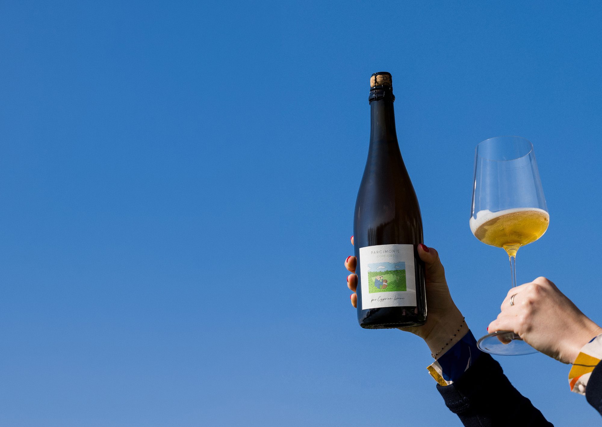 A hand raising a bottle of Parcimonie cidre against a deep blue summer sky, a glass of sparkling cider alongside