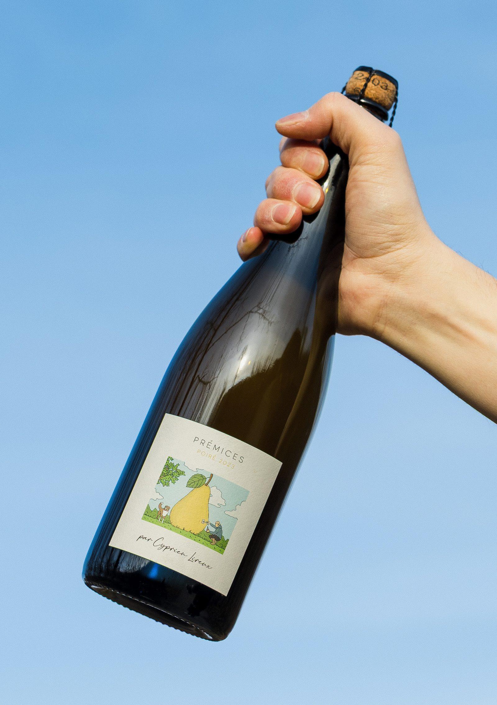 A hand holding a bottle of Prémices poiré aloft against a clear blue sky