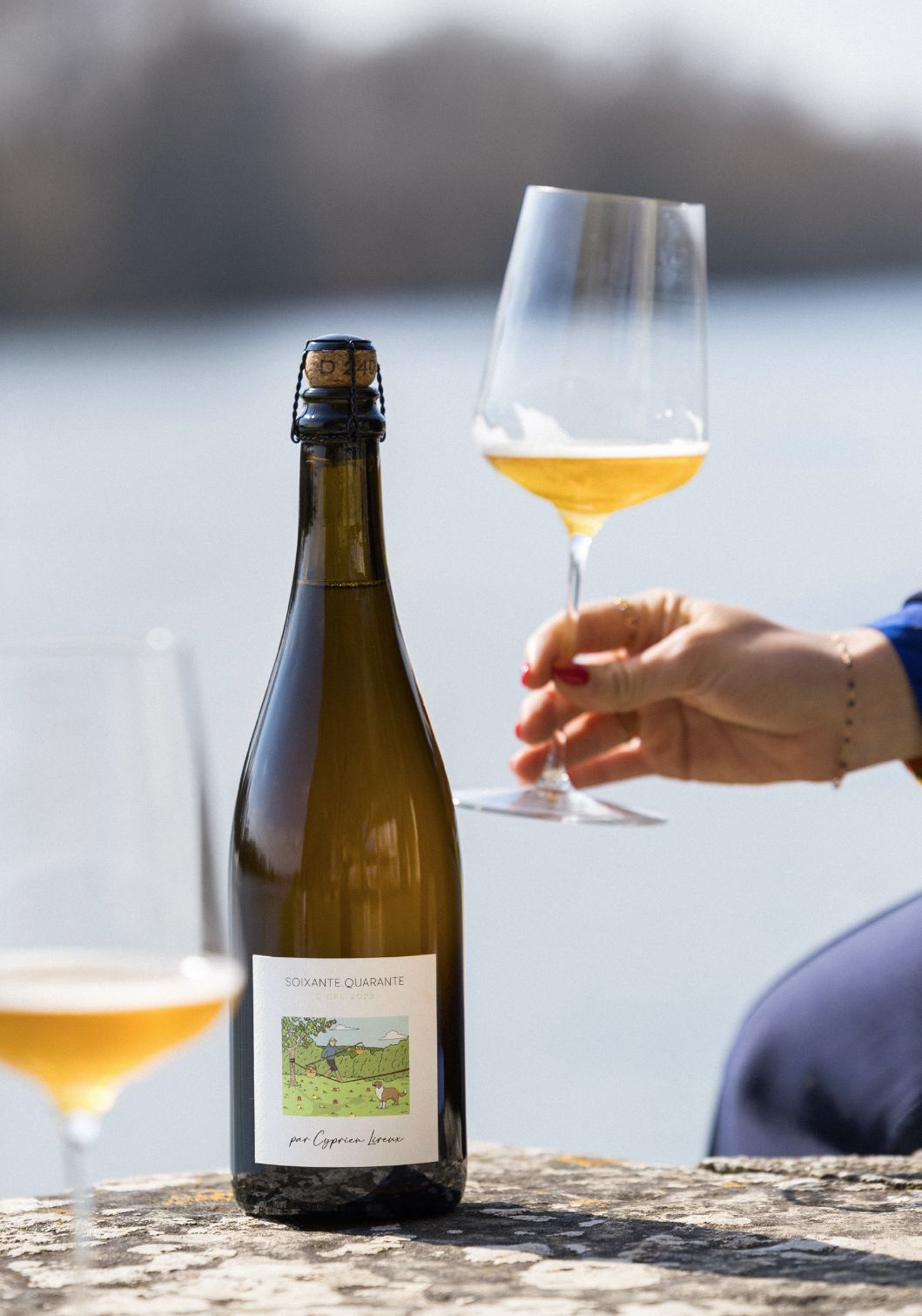 A bottle of Soixante Quarante standing on a lichen-covered stone wall by the water, a hand raising a glass of pale gold cider beside it