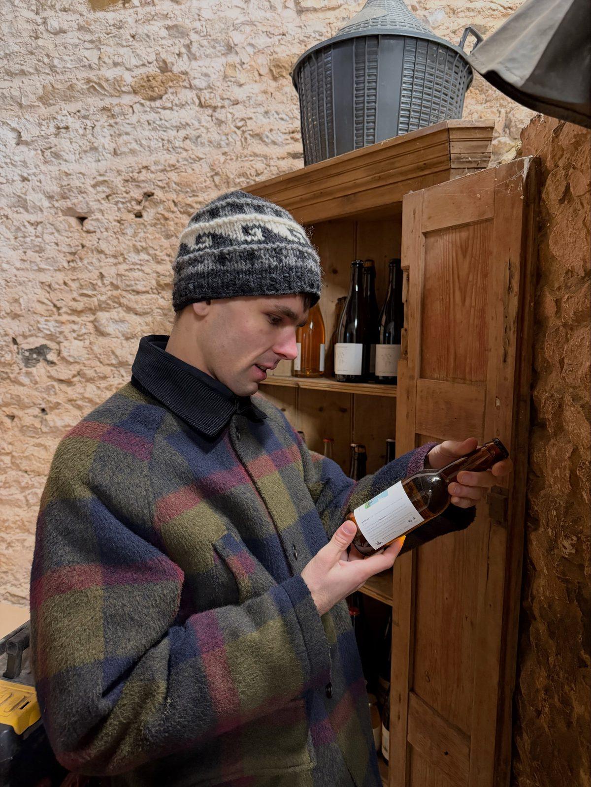 Jack Laycock, founder of Cidermugs, examining a bottle beside a wooden cabinet in the cellar at Domaine Cyprien Lireux