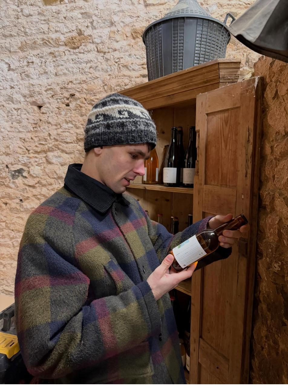 Jack examining a bottle of cider in a producer's cellar, wearing a checkered wool jacket and patterned knit beanie, with shelved bottles behind