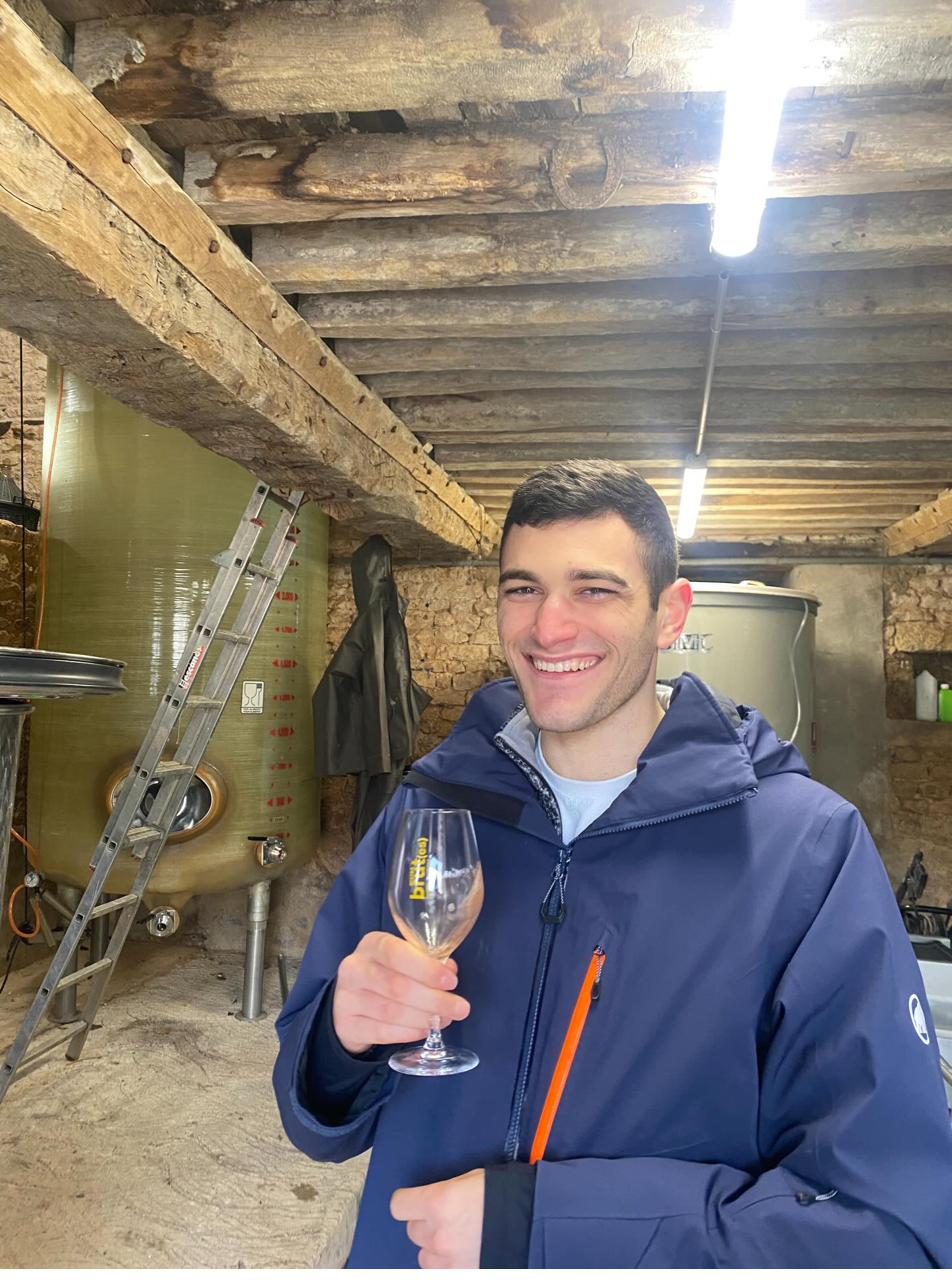 Yiannis smiling and raising a tasting glass in a cider barn, with a fibreglass fermentation tank and ladder behind