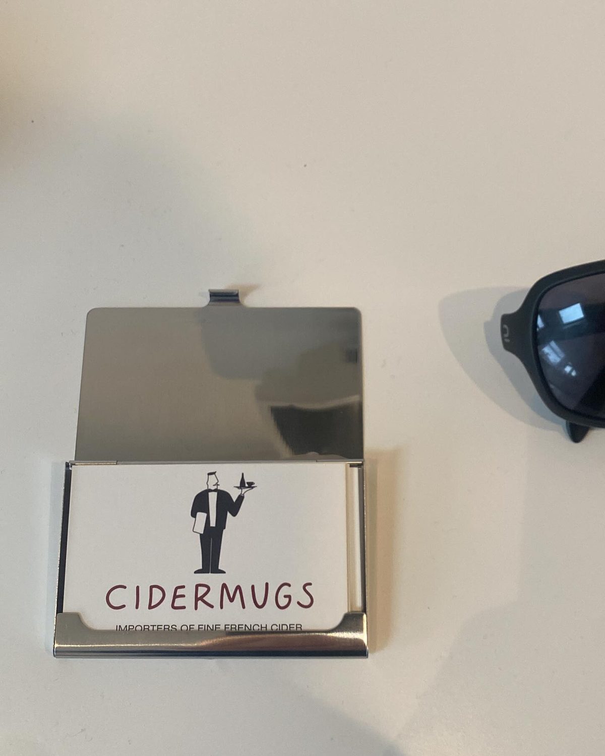 A polished metal business card holder, open to reveal a Cidermugs card showing the waiter logo and 'IMPORTERS OF FINE FRENCH CIDER'