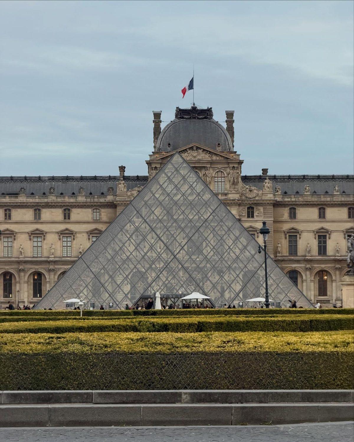 The glass pyramid at the Louvre against the museum's stone facade, a French flag flying above
