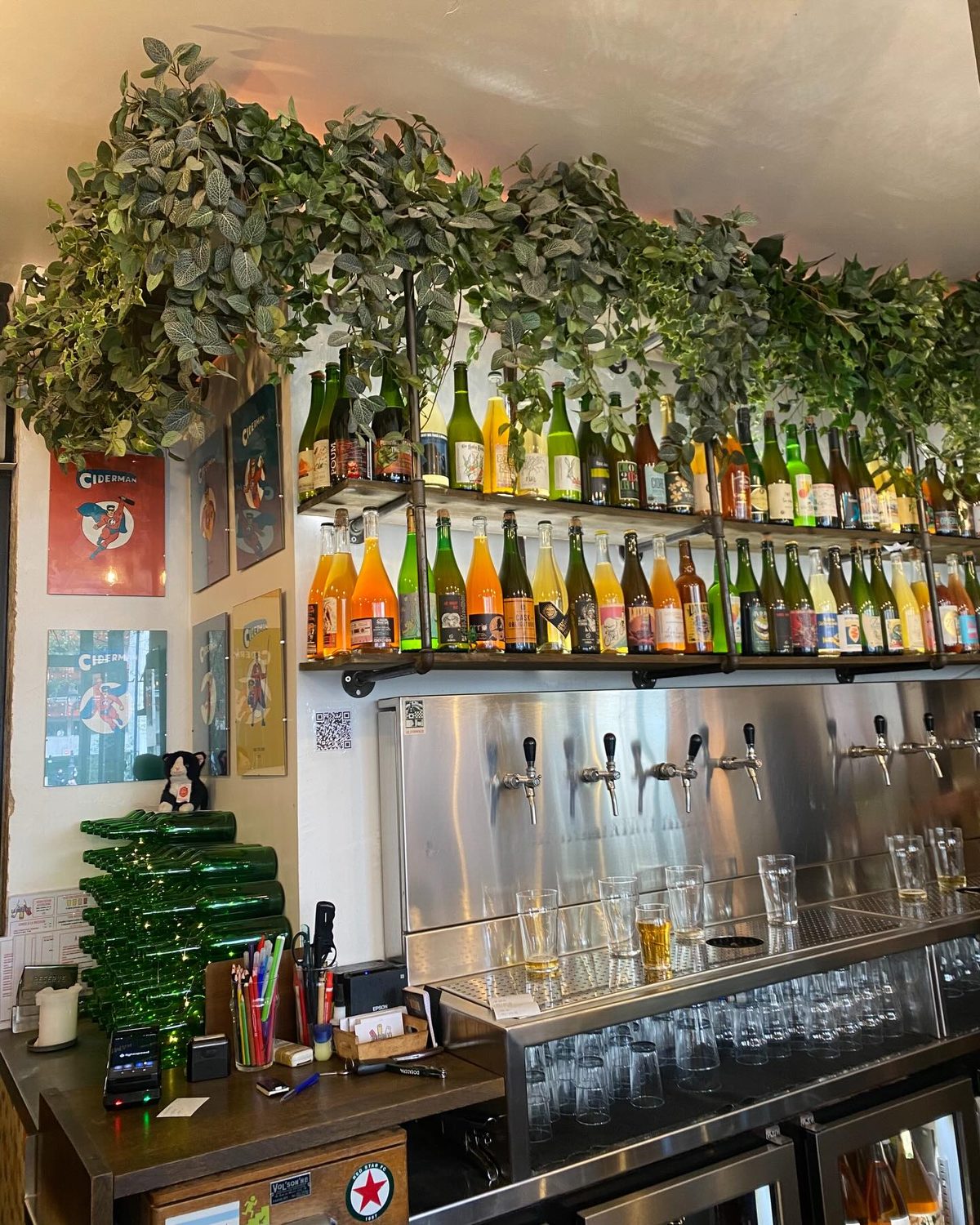 The bar at La Cidrerie Paris - rows of natural cider bottles on shelves beneath trailing greenery, with cider taps and tasting glasses lined up below