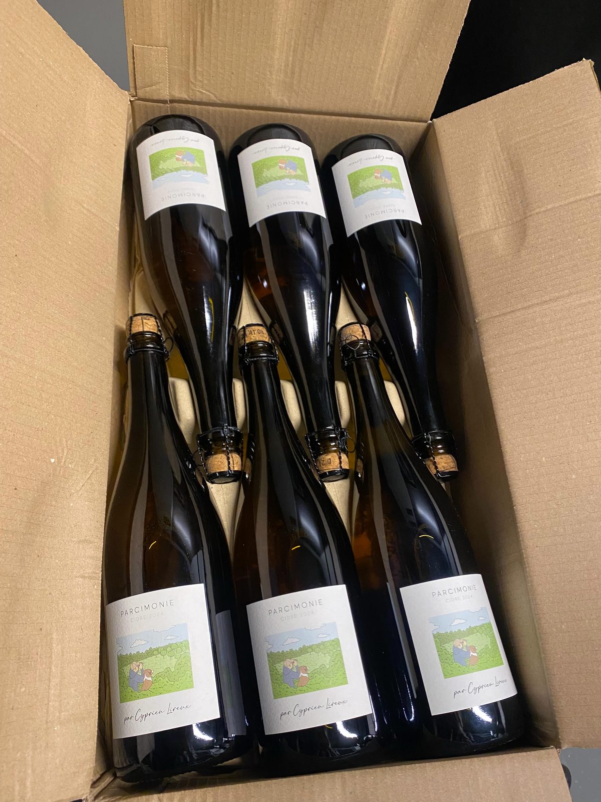 Six bottles of Parcimonie 2024 cidre packed in a cardboard case, the three at the front showing labels with Cyprien Lireux's green pastoral illustration of a couple in an orchard