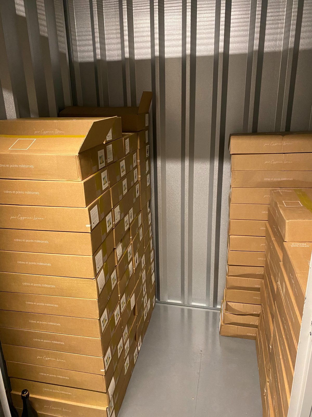 Tall stacks of brown Cyprien Lireux cider cases inside a metal storage unit, the cases labelled 'cidres et poirés millésimés'
