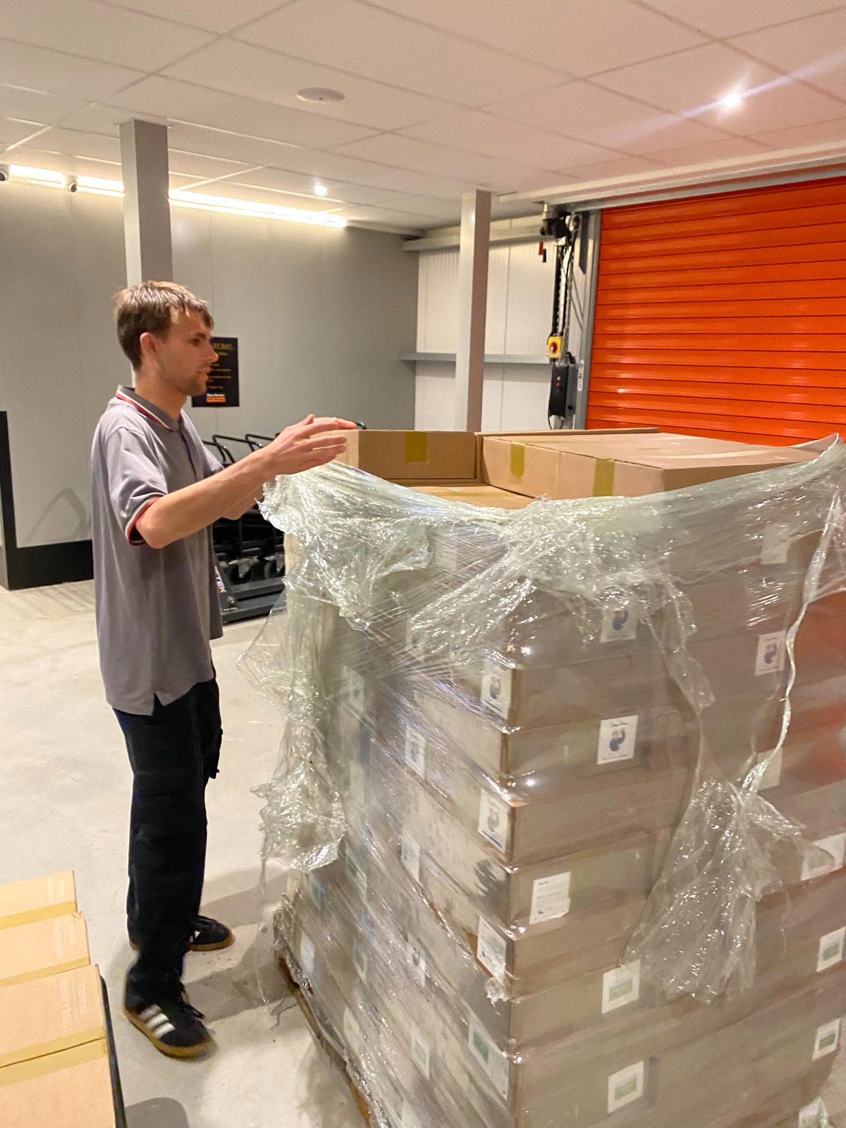 Jack at a UK warehouse loading bay, gesturing at a shrink-wrapped pallet of Cidermugs cases just delivered, with an orange roller door behind