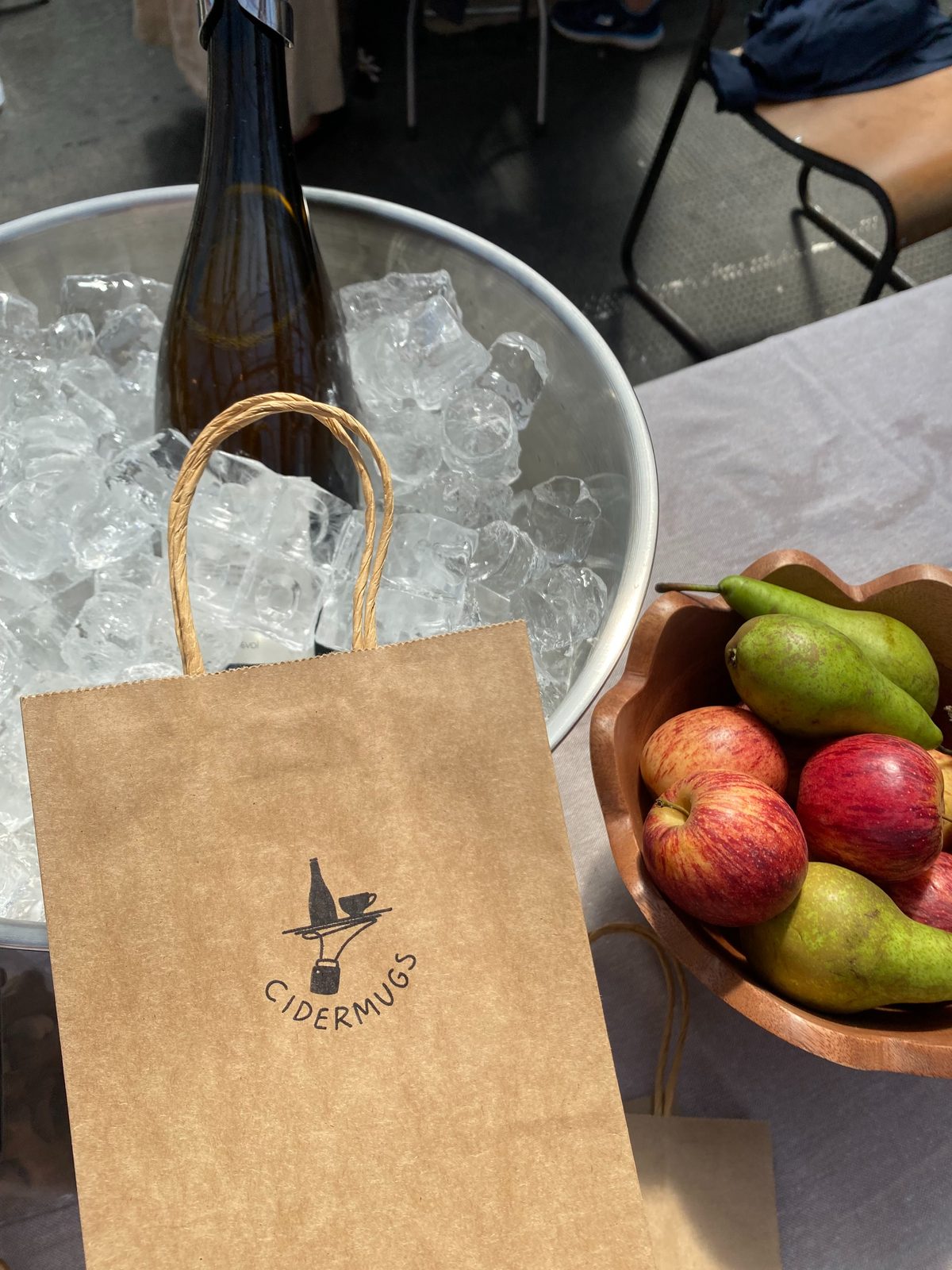 An ice bucket, a Cidermugs paper bag and a bowl of apples and pears on the stall table