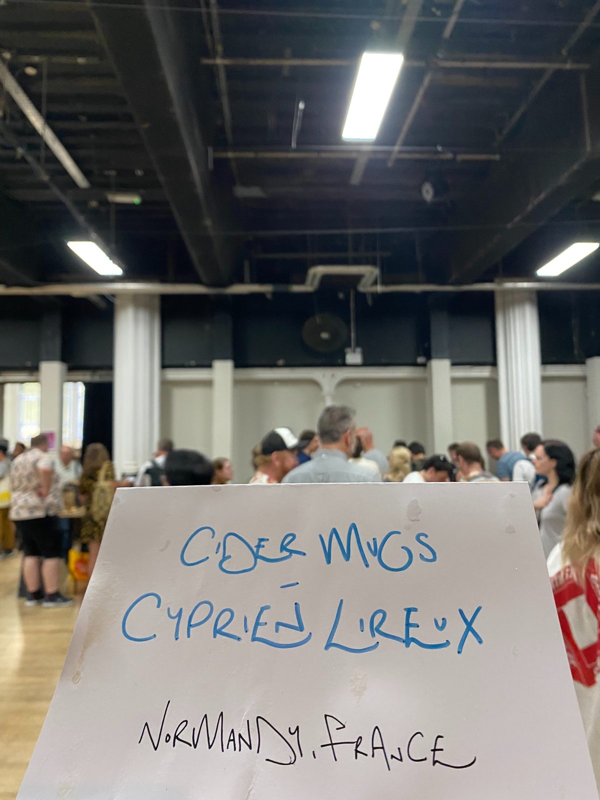 A handwritten table sign reading 'Cidermugs, Cyprien Lireux, Normandy France', held up in the busy salon hall