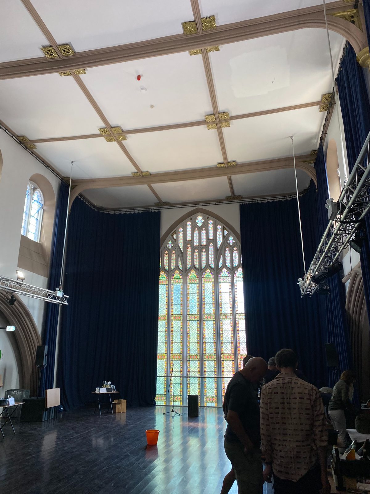 A grand hall with a towering arched window and deep blue curtains, set for the weekend's gathering