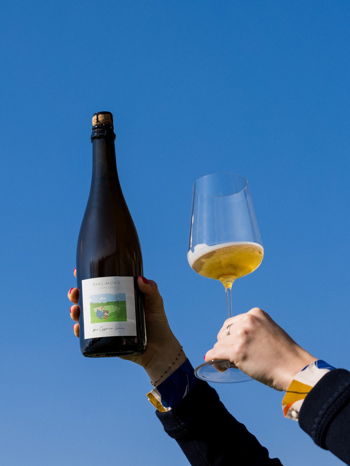 A bottle of Parcimonie and a brimming glass raised in a toast against a clear blue sky