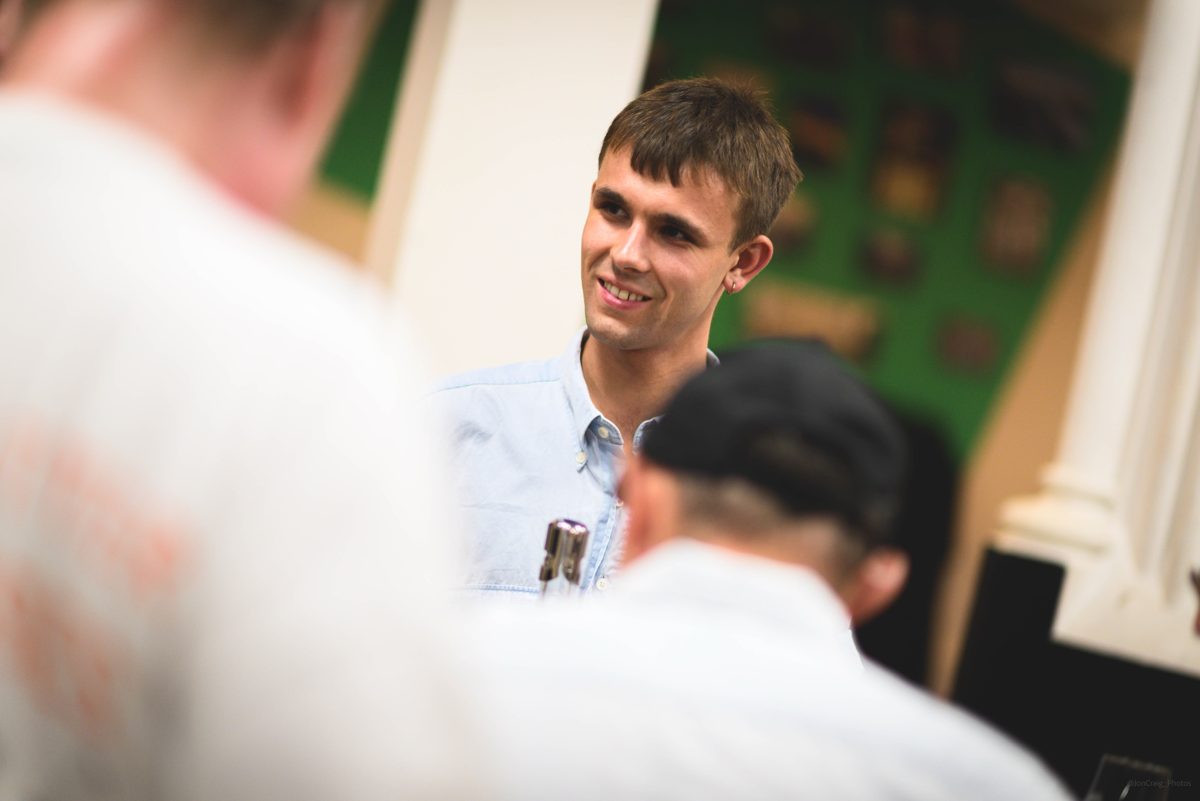 Jack in a pale blue shirt, smiling as he talks with tasters across the table at Bristol Cider Salon, a bottle cap tucked in his shirt pocket